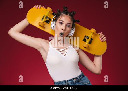 Foto di un scioccato emotivo punk giovane ragazza adolescente ascoltare musica con le cuffie isolate su Borgogna rosso sfondo parete azienda skateboard. Foto Stock