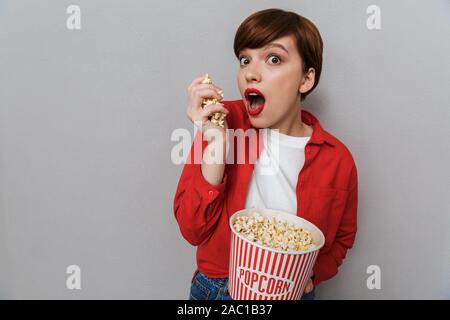 Immagine della donna sorpresa tenendo la carta benna e mangiare popcorn isolate su uno sfondo grigio Foto Stock