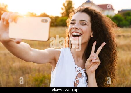 Foto di ricci eccitato donna prendendo selfie foto sul cellulare e gestualità segno di pace mentre passeggiate in campagna durante la giornata di sole Foto Stock