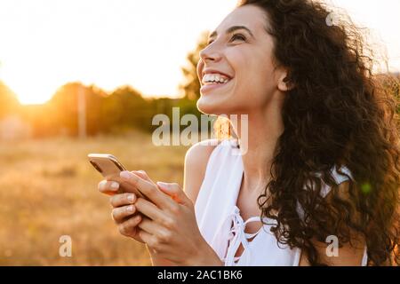 Foto di stupiti giovane donna digitazione sul cellulare e sorridere mentre passeggiate in campagna durante la giornata di sole Foto Stock