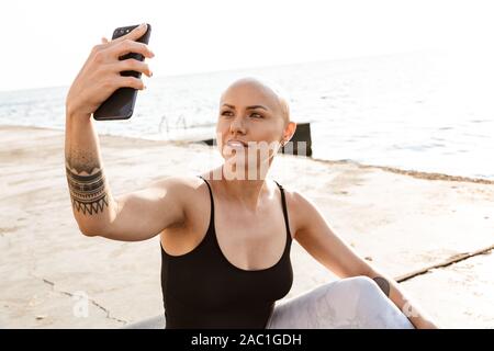 Immagine della lieta bald donna in abbigliamento sportivo tenendo selfie foto sul cellulare mentre è seduto vicino al mare nella mattina di sole Foto Stock