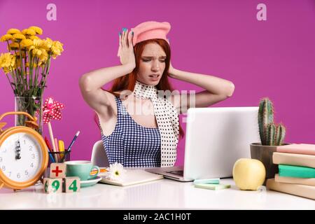 Immagine del sottolineato teen ragazza lo studio sul computer portatile mentre è seduto alla scrivania isolate su sfondo viola Foto Stock