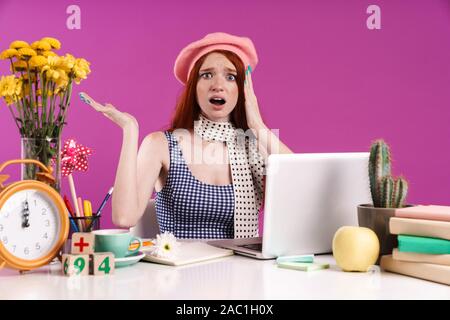 Immagine di frustrati teen ragazza lo studio sul computer portatile mentre è seduto alla scrivania isolate su sfondo viola Foto Stock