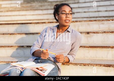 Attraente giovane donna africana di indossare un abbigliamento informale studiare seduti sui gradini esterni, indossa gli auricolari wireless Foto Stock