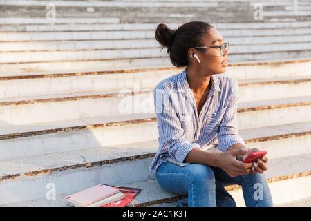 Attraente giovane donna africana di indossare un abbigliamento informale l'ascolto di musica con cuffie e tenere il telefono cellulare mentre è seduto sui gradini esterni Foto Stock