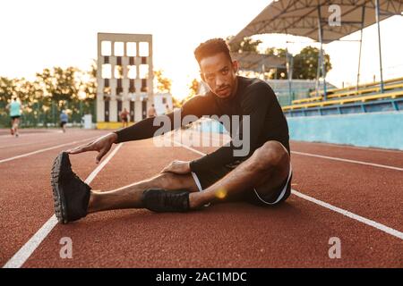 Immagine di concentrato di African American uomo stretching il suo corpo mentre è seduto a terra di sport all'aperto Foto Stock