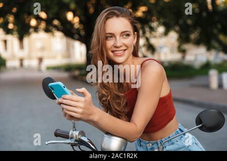 Bella sorridente ragazza casual da indossare abbigliamento estivo seduto su uno scooter all'aperto presso le strade della città, utilizzando il telefono cellulare Foto Stock