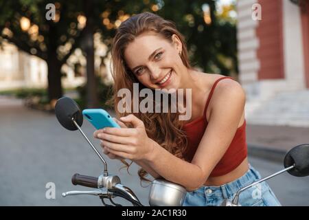 Bella sorridente ragazza casual da indossare abbigliamento estivo seduto su uno scooter all'aperto presso le strade della città, utilizzando il telefono cellulare Foto Stock