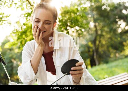 Bella sorridente ragazza casual da indossare abbigliamento estivo seduto su uno scooter all'aperto presso le strade della città, guardando lo specchio Foto Stock