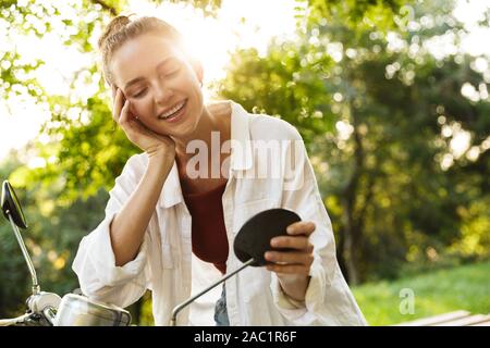 Bella sorridente ragazza casual da indossare abbigliamento estivo seduto su uno scooter all'aperto presso le strade della città, guardando lo specchio Foto Stock