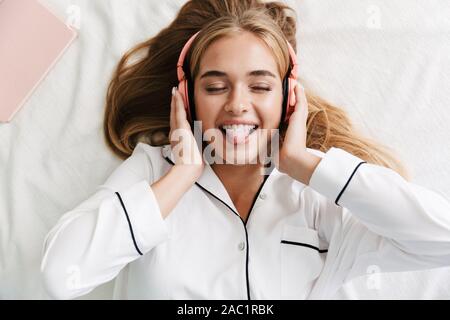 Immagine di gioiosa giovane ragazza nelle cuffie sporgenti dalla sua lingua mentre giaceva a letto a casa Foto Stock