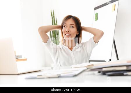Attraente sorridente giovane imprenditrice asiatici lavorare mentre è seduto alla scrivania in ufficio, rilassante sulla sedia Foto Stock