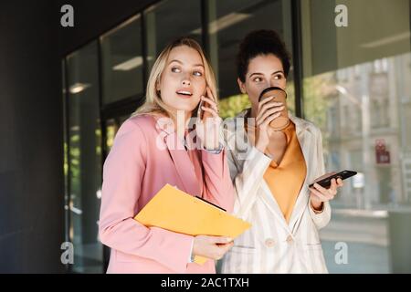 Due belle sorridenti giovani imprenditrici passeggiate all'aperto presso le strade della città, parlare mentre si beve il caffè e utilizzando i telefoni cellulari Foto Stock