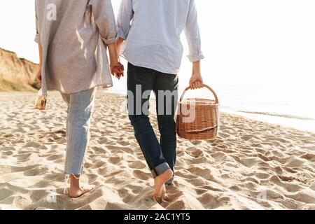 Immagine ritagliata vista posteriore di una giovane coppia di trascorrere del tempo in spiaggia, portando un cestino picnic mentre passeggiate in spiaggia Foto Stock