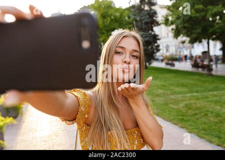 Bella sorridente ragazza bionda sta prendendo un selfie con telefono cellulare mentre in piedi all'aperto, invio di kiss Foto Stock