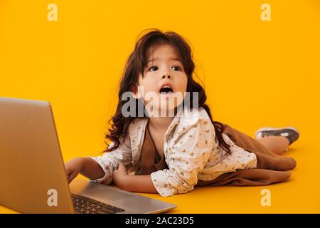 Immagine di stupito bella ragazza digitando su laptop mentre giaceva sul pavimento isolato su sfondo giallo Foto Stock