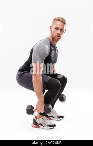 Fiducioso attraente giovane sportivo muscolare in piedi isolato su sfondo bianco, il sollevamento di un carico pesante barbells, accovacciata Foto Stock