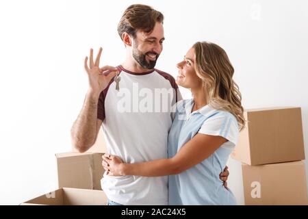 Foto della coppia sorridente in abbigliamento casual abbracciando vicino a scatole di cartone e tenendo premuto i tasti isolate su muro bianco Foto Stock