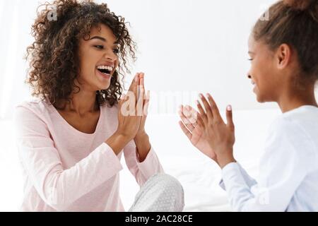 Immagine di gioiosa americano africano la donna e la sua piccola figlia sorridente e battendo le mani sul divano di casa Foto Stock