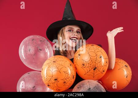 Allegro bambina indossa Halloween costume strega in piedi isolato su sfondo rosso, tenendo mazzetto di palloncini Foto Stock