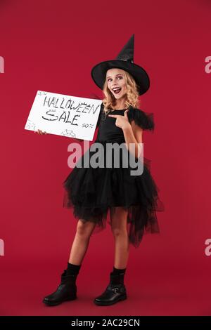 Immagine positiva di piccola ragazza strega in carnevale costume di halloween isolato sulla parete rossa sullo sfondo azienda copyspace vuota con la vendita di testo. Foto Stock