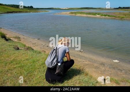 Knokke-Heist, Belgio, 29 luglio 2019 Parco Naturale Zwin, donna bionda con occhiali da sole seduto sul bordo dell'acqua Foto Stock