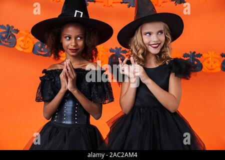 Immagine di belle ragazze multinazionale in nero costumi di halloween sorridente e di stare insieme isolate su orange parete di zucca Foto Stock