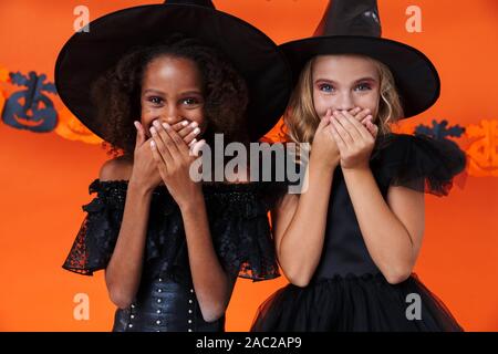 Immagine della cute ragazze multinazionale in nero costumi di halloween a ridere e coprire le loro bocche isolate su orange parete di zucca Foto Stock