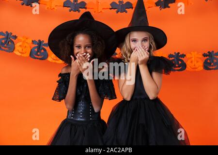 Immagine di pretty multinazionale di ragazze in nero costumi di halloween a ridere e coprire le loro bocche isolate su orange parete di zucca Foto Stock