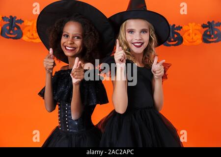 Immagine di gioiosa multinazionale di ragazze in nero costumi di halloween cercando e dita di puntamento in telecamera isolate su orange parete di zucca Foto Stock