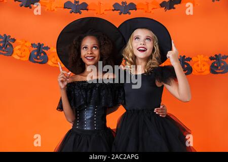 Immagine della multinazionale sorridenti ragazze in nero costumi di halloween abbracciando e denti rivolti verso l'alto isolato su orange parete di zucca Foto Stock