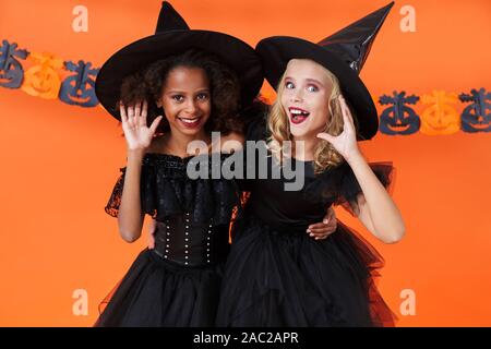 Immagine di lieti multinazionale di ragazze in nero costumi di halloween e abbraccia l'ascolto isolate su orange parete di zucca Foto Stock