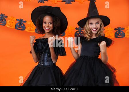 Immagine di felice multinazionale di ragazze in nero costumi di halloween sorridente con lanci pugni isolate su orange parete di zucca Foto Stock