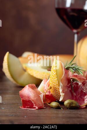 Bicchiere di vino rosso e spuntini mediterranei. Il Prosciutto e melone, capperi e rosmarino su un vecchio tavolo di legno, copia dello spazio. Foto Stock