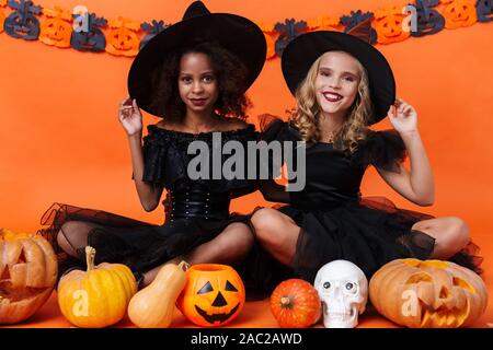 Immagine di felice multinazionale di ragazze in nero costumi di halloween sorridere mentre seduto con zucche e toy cranio isolate su orange parete di zucca Foto Stock