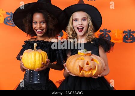 Immagine di ridere multinazionale di ragazze in nero costumi di halloween zucche di contenimento isolato sulla parete arancione Foto Stock