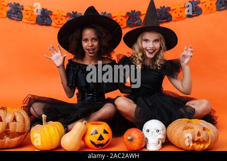 Immagine di felice multinazionale di ragazze in nero costumi di halloween di graffiare mentre è seduto con zucche e toy cranio isolate su orange parete di zucca Foto Stock