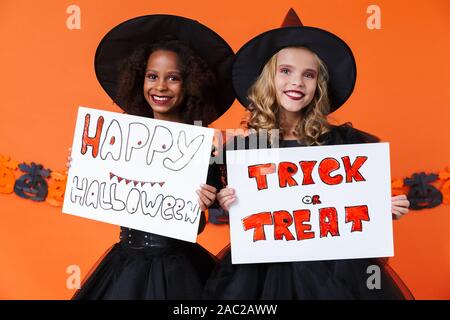 Immagine della multinazionale sorridenti ragazze in nero costumi di halloween holding placards isolate su orange parete di zucca Foto Stock