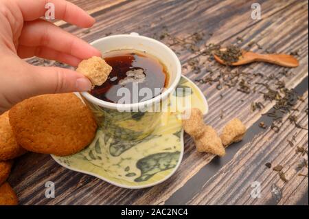 La mano della ragazza mette un pezzo di zucchero di canna in una tazza di tè nero, fiocchi d'avena cookies, foglie di tè e zucchero di canna su uno sfondo di legno. Close up Foto Stock