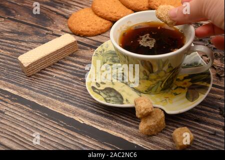 La mano della ragazza mette un pezzo di zucchero di canna in una tazza di tè nero, fiocchi d'avena cookies, foglie di tè, waffle, lo zucchero di canna su uno sfondo di legno. Close up Foto Stock