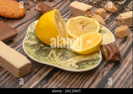 Fette di limone su un piattino, pezzi di zucchero di canna, fiocchi d'avena biscotti, cialde, pezzi di cioccolato su uno sfondo di legno. Close up Foto Stock