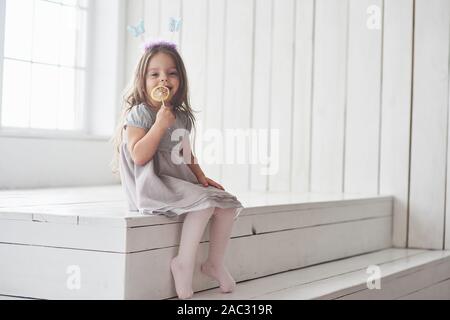 Seduta sulle scale. Graziosa bambina di mangiare la candela gialla con farfalle giocattolo sulla sua testa nella camera bianca Foto Stock