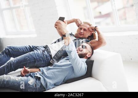 Due eleganti giovani amici la posa sul divano e tenendo selfie. Ragazzo biondo mostra bicipite Foto Stock