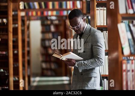 Smart cercando african american businessman in bicchieri permanente e la lettura di un libro in biblioteca Foto Stock