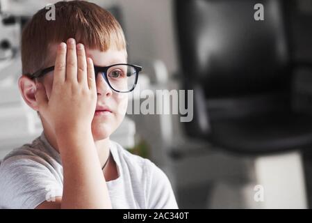 Vista frontale. Chiudere occhio destro con la mano. Little Boy controllando la sua visione con nuovi occhiali nero Foto Stock