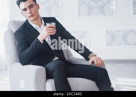 Imprenditore guarda nella telecamera. Giovane ragazzo elegante in abito nero seduto sulla sedia bianca e detiene il vetro con alcol Foto Stock