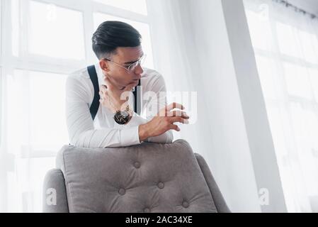 Guarda nel bicchiere di whiskey. Giovane uomo bello in abbigliamento classico e eyewears appoggiata sulla sedia mentre in piedi contro finestre bianche Foto Stock