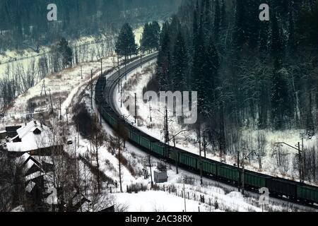 Treno merci si muove attraverso la foresta di conifere lungo il fiume congelato Foto Stock