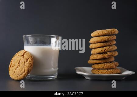 Un cookie di zenzero poggia su un bicchiere di latte accanto a una pila di biscotti allo zenzero su una piastra bianca e uno sfondo nero; spazio di copia Foto Stock