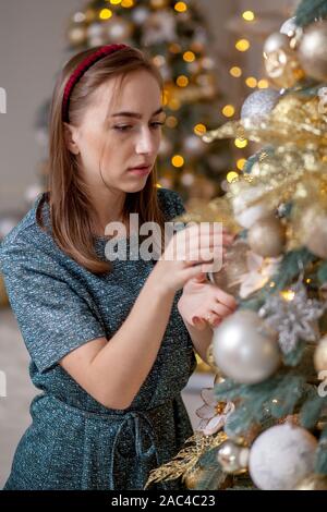 Bella ragazza decorare albero di Natale. Una giovane donna sorridente prepara un albero di Natale per la vacanza. Lussureggiante verde albero di Natale con palline dorate Foto Stock